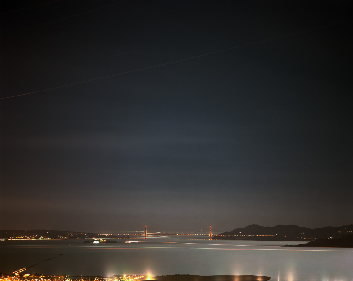 Long-exposure color photograph of the distant Golden Gate Bridge lit up at night with city skyline lights and moving light trails of boats in the foreground