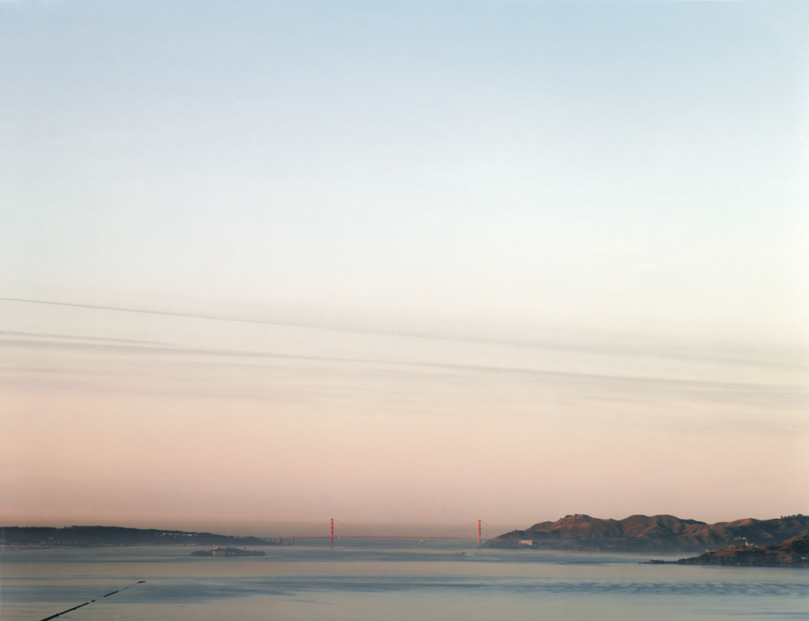 Color photograph of the distant Golden Gate Bridge under a clear pale blue and pink sky