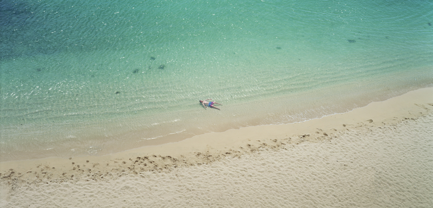 Color photograph of a person floating on their back in shallow blue-green water near the shore