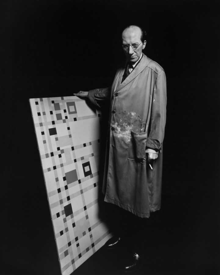 Black-and-white frontal portrait of a wax figure of a man in a long artist's smock with an abstract painting of small squares propped upright in one hand