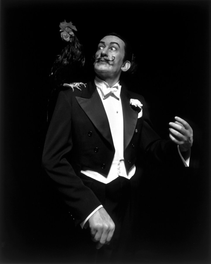 Black-and-white frontal portrait of a wax figure of a mustached man in a white-tie tuxedo looking in surprise at a rooster on his shoulder