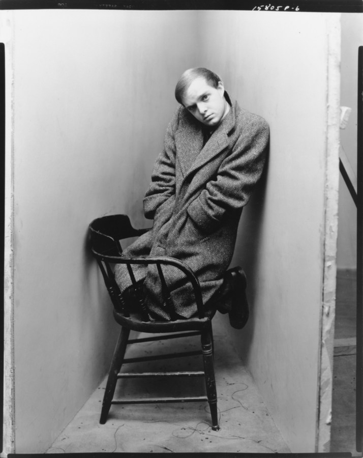 Black-and-white photograph of a man in a long coat kneeling on a wooden chair
