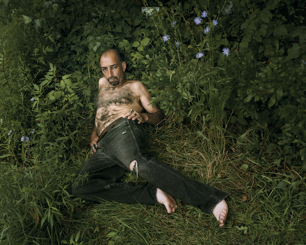 Color photograph of a shirtless man reclining in a patch of long grass
