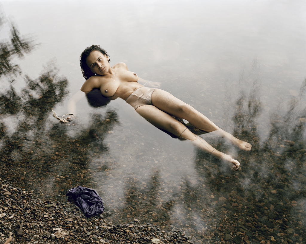 Color photograph of a shirtless woman reclining on her back in shallow water next to a pebbled shore