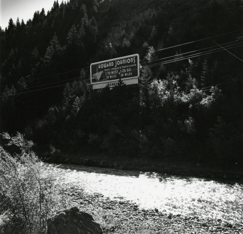 A black and white photograph of a billboard advertising Howard Johnson's against pine trees and a river in the foreground.