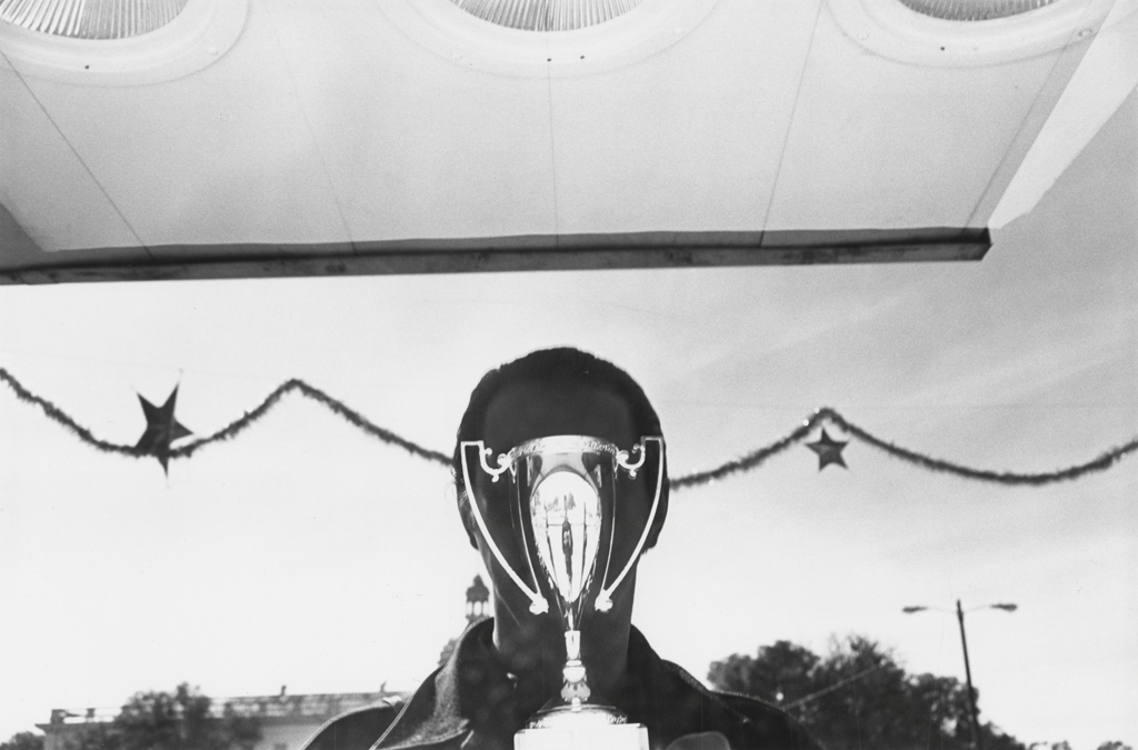 A black and white photograph of the reflection of the artist over a shining trophy in a store window