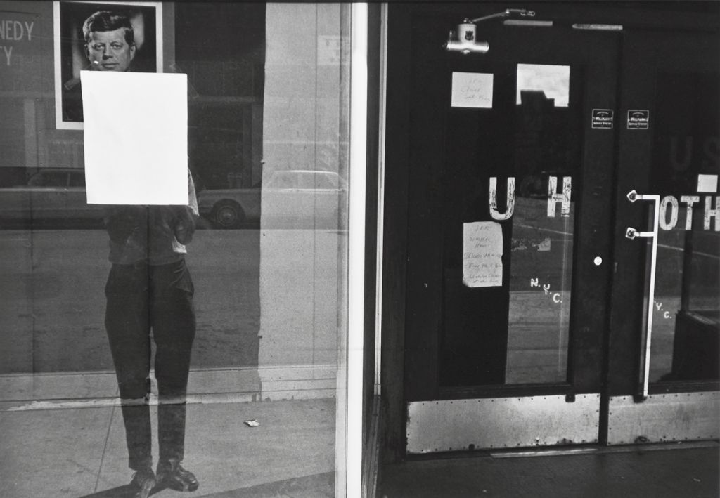 A black and white photograph of a self portrait of the artist reflected in a shop window