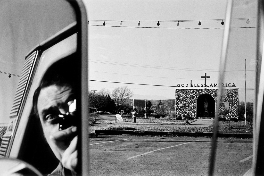 A black and white photograph of a self portrait of the artist reflected in the side mirror of a car with a church in the right of the frame