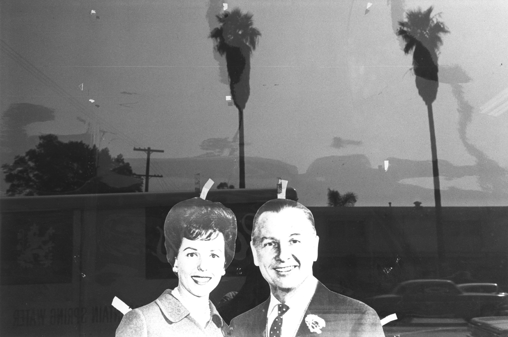 Black-and-white photograph of cut outs of a man and woman taped in a window with palm trees in the reflection