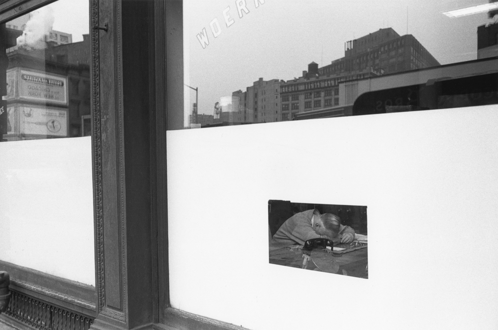 Black-and-white photograph of a man with laying his head on a table through the window of a storefront