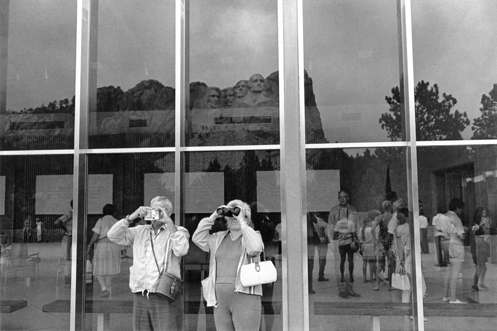 Black-and-white photograph of a man and woman looking out of the frame with binoculars with reflections in the glass wall behind them