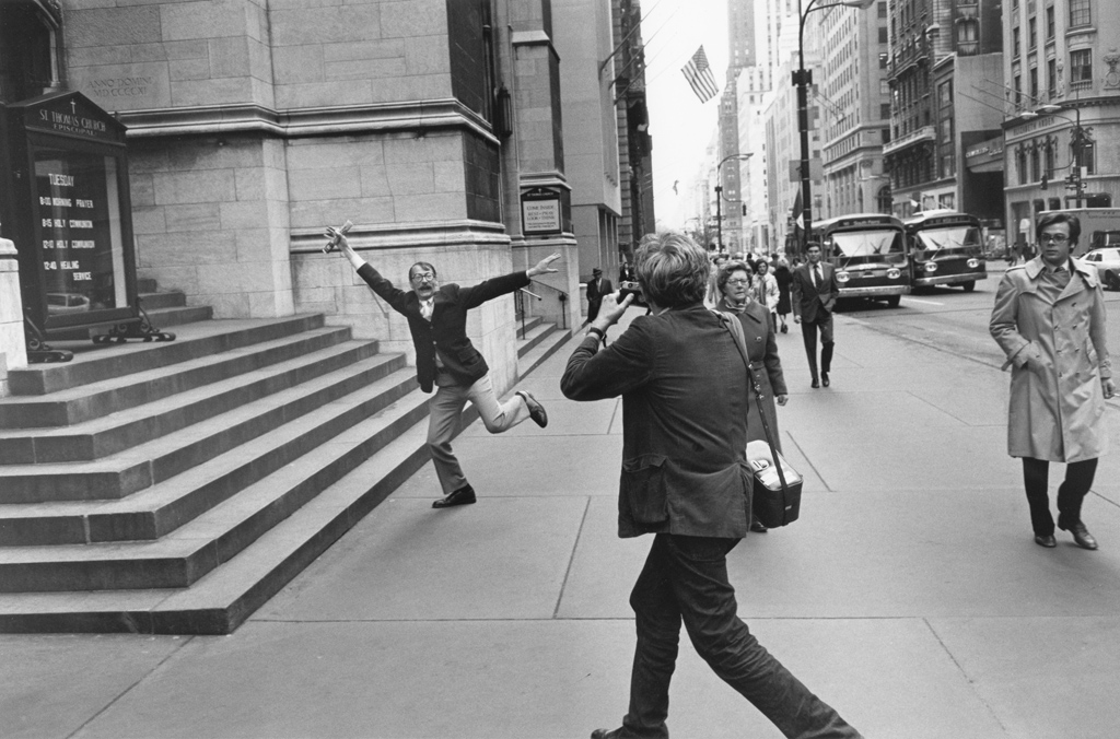 Black-and-white photograph of a man taking a photograph of a man doing a jumping motion on a city sidewalk