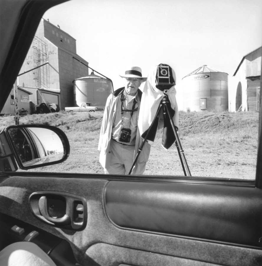 Black and white photograph out the window of a car of a man with a camera on a tripod