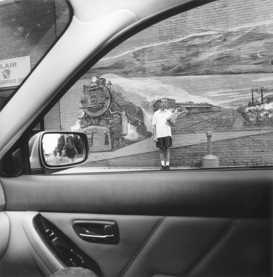 Black and white photo looking out the window of a car at a person standing in front of a mural