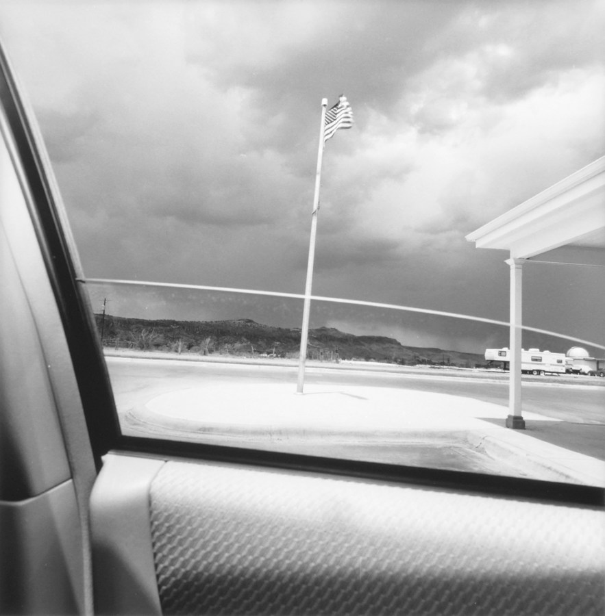 Black and white photograph out the window of a car of a flag pole with American Flag and overcast sky