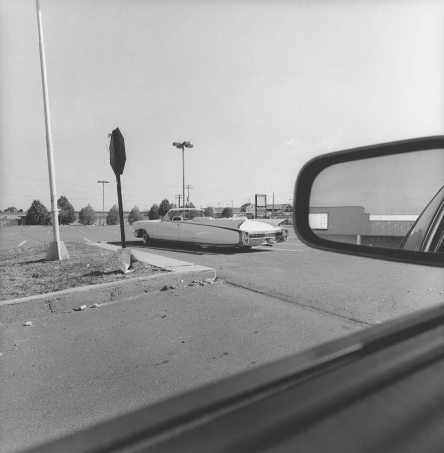 Black and white photograph out the window of a car of a 1950s convertible parked in an empty parking lot