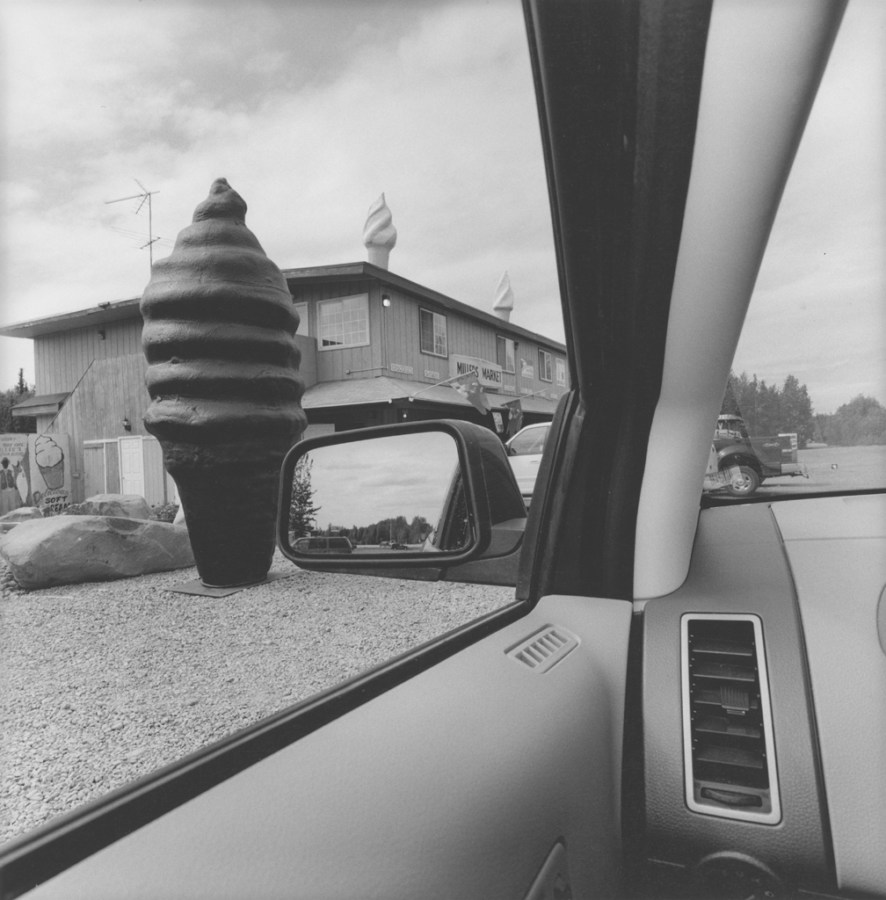 Black and white photograph out the window of a car of a large fake ice cream cone roadside attraction