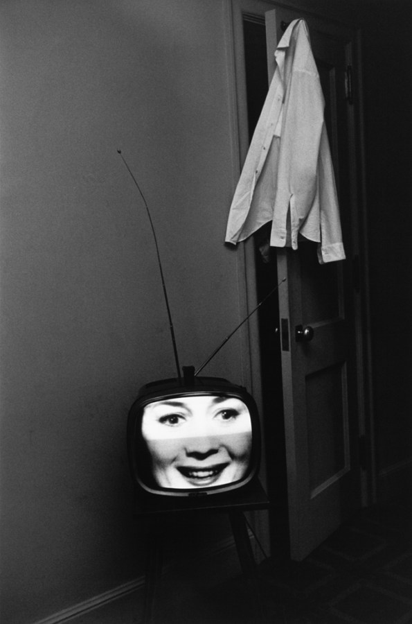 Black-and-white photograph of a television with a laughing face on screen and a shirt hanging from an open door
