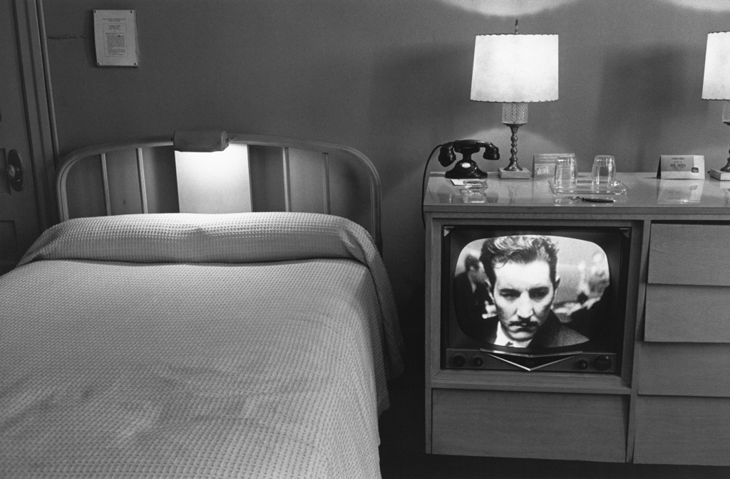 Black-and-white photograph of a neatly made bed and a television with a mans face on screen