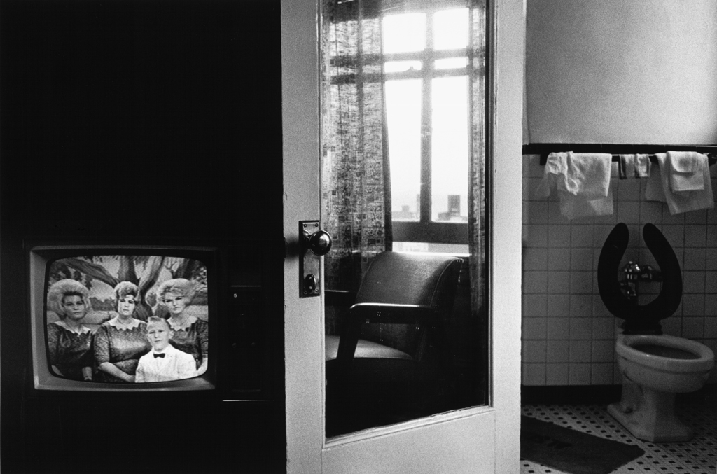 Black-and-white photograph of a television with figures on screen, a reflection in a door mirror, and a toilet