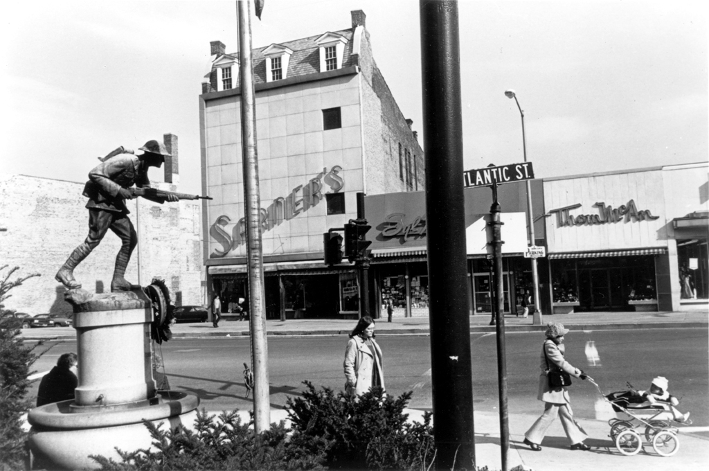 Black and white photograph of a monument of a man holding a gun and people walking in the direction the gun is pointed