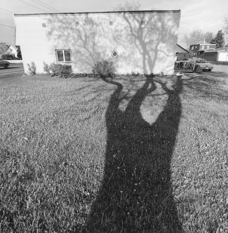 A black and white photograph of a self portrait of the shadow of a tree against a house