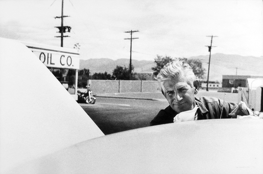 Black-and-white photograph of a man with white hair peering over the roof of a car in a parking lot