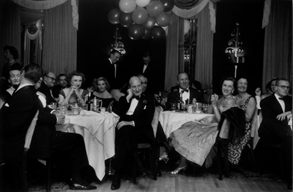 Black-and-white photograph of a party setting with men and women in formal dress seated at round tables