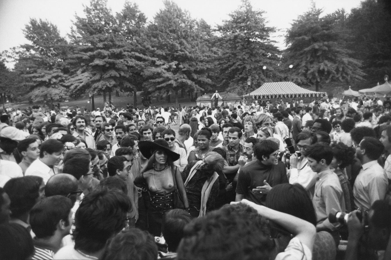 Black-and-white-photograph of a bare-breasted woman wearing a large hat in a crowd of people in a park