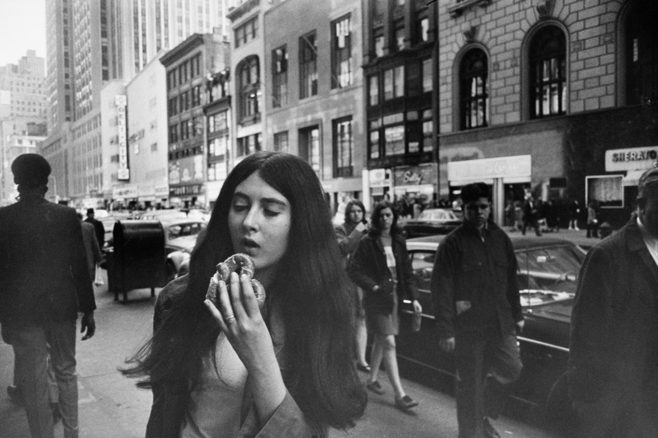 Black-and-white photograph of a woman on a city sidewalk holding a pretzel to her mouth