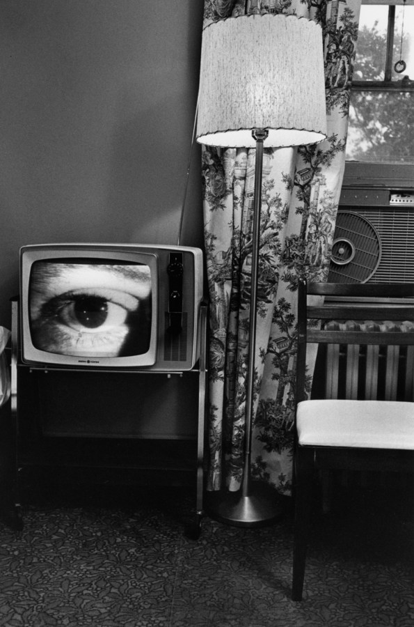 Black-and-white photograph of room with a lamp a chair and a television with a close up of an eye on screen