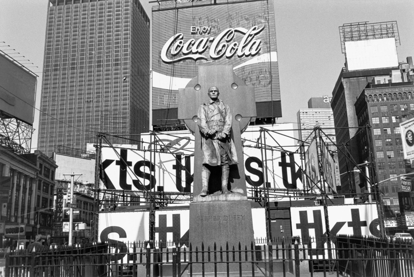 Black-and-white photograph of a statue with a Coca-Cola sign, billboards, and skyscrapers in the background