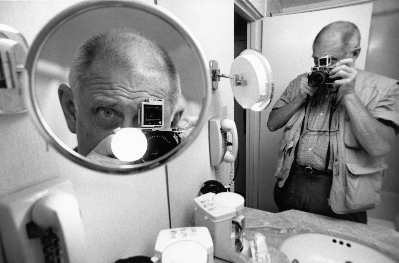 A black and white photograph of a self portrait of the artist in the reflection of two different bathroom mirrors