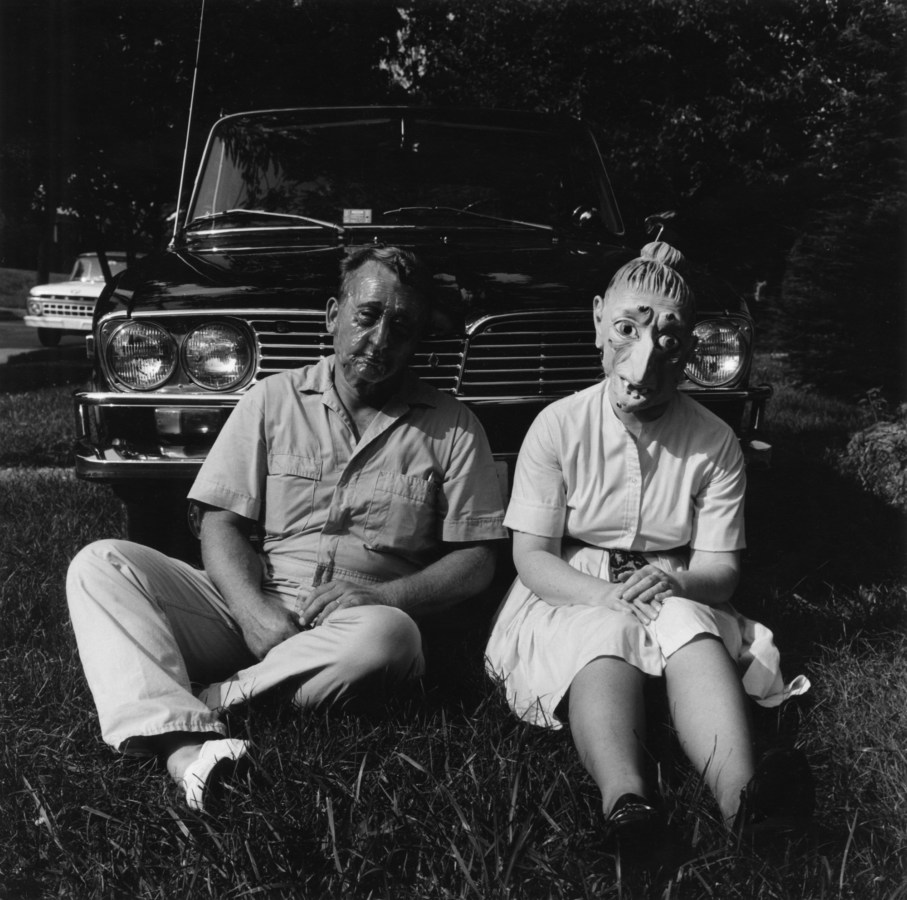 Black-and-white photograph of a couple wearing rubber masks seated in the grass in front of a car