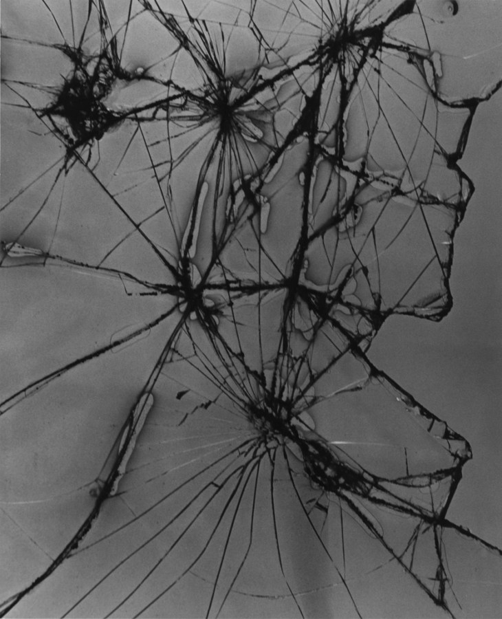Black-and-white photograph of a cracked pane of glass with spider-like converging fracture patterns