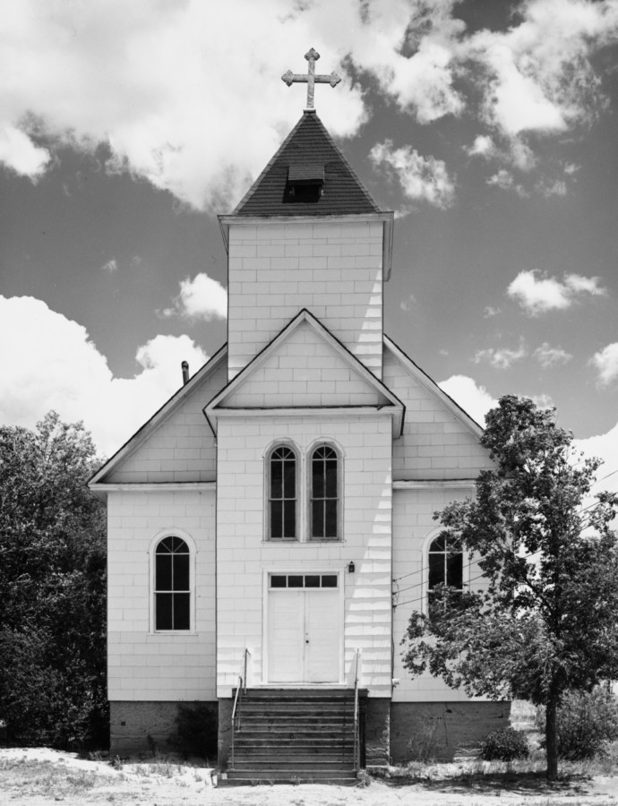 A vertical black and white photograph of an old wooden church with trees at each side and a cloudy sky.