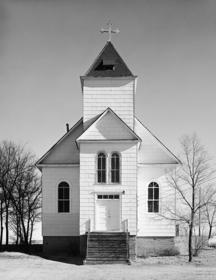 A vertical black and white photograph of an old wooden church with trees at each side and a clear bright sky.