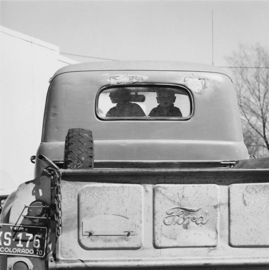 A black and white photograph of two children looking out the rear window of a beat up early 1950's Ford pick up truck.