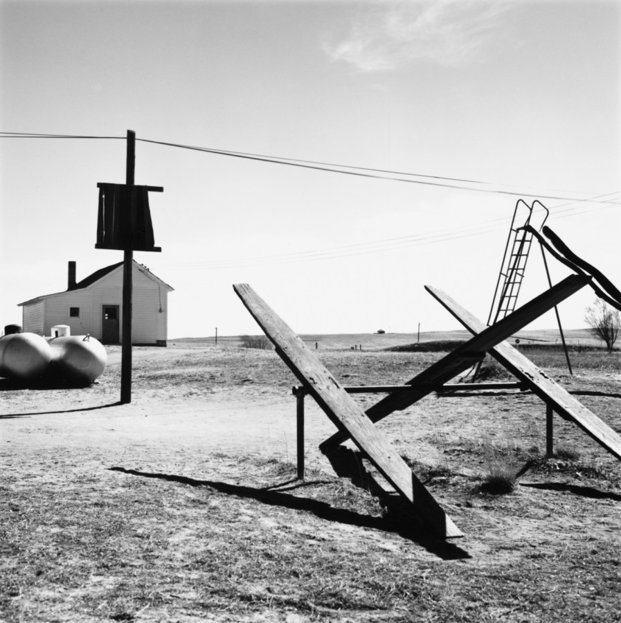 A black and white photograph of a playground with teeter totters, a slide, and a telephone pole with wires. A wooden structure in the background against a bright clear sky.