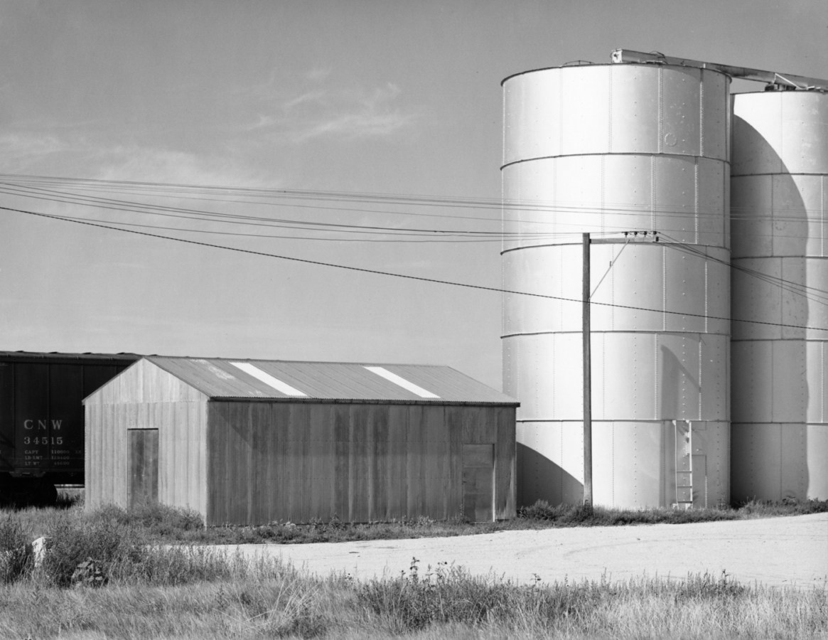 A black and white photograph of a train car, a windowless structure, and silos against a brightly lit sky and telephone wires.