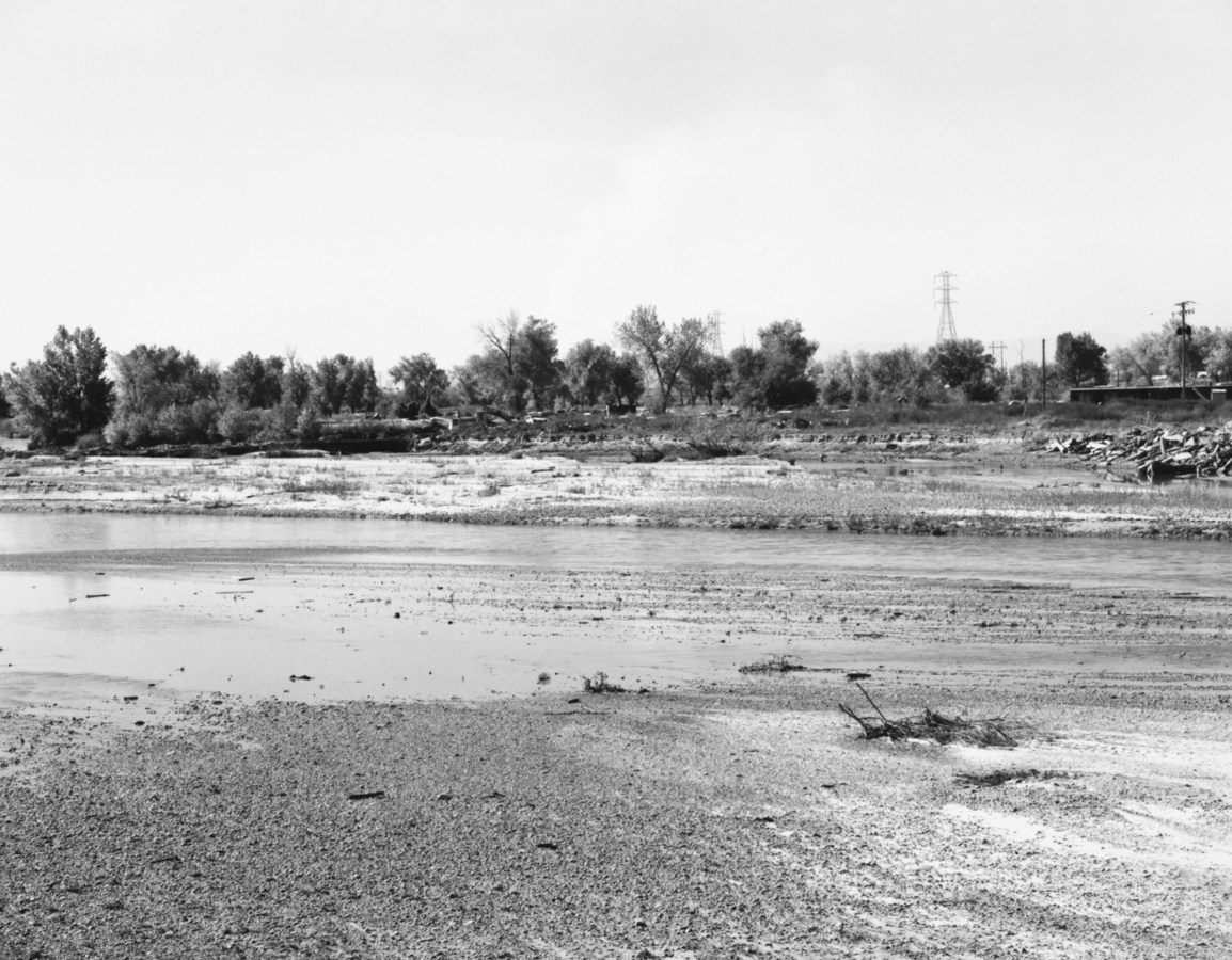 A black and white photograph of a river with trees and telephone wires in the background.