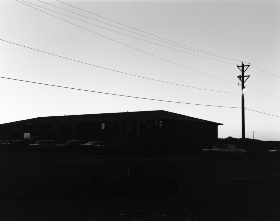 A black and white photograph of an apartment building and parked cars in shadow against a clear sky.