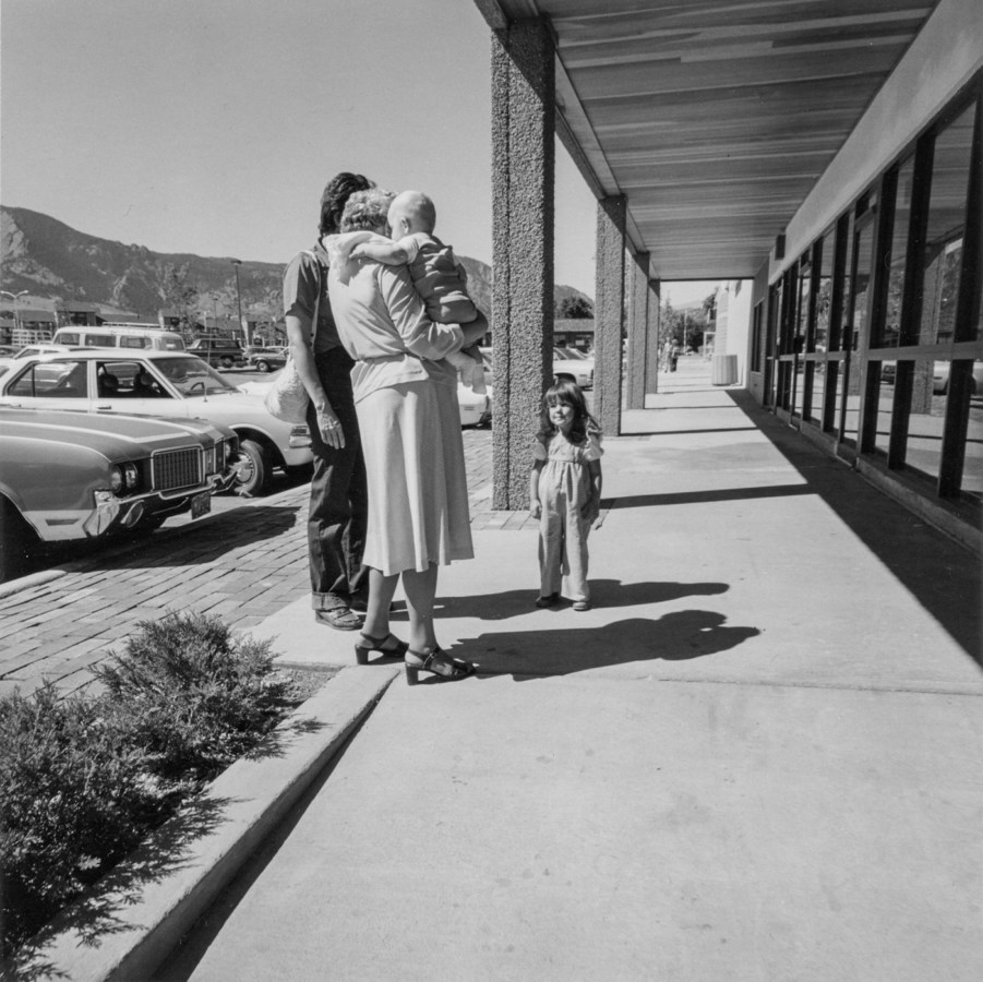 Black-and-white photograph of a man and woman holding a child with their backs to the camera, another child facing the camera. Parked cars and storefronts are in the background.