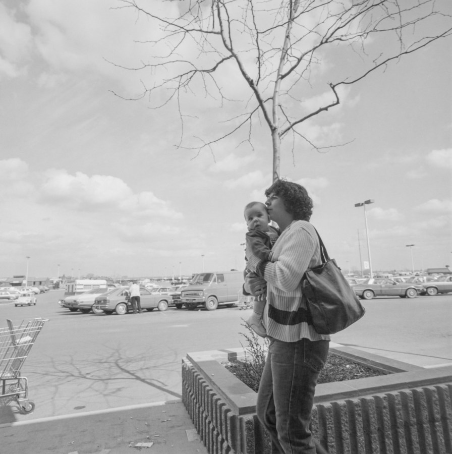 Black-and-white photograph of a woman holding a baby, in the background is a tree with no leaves in the center, a shopping cart to the left, and a parking lot filled with cars