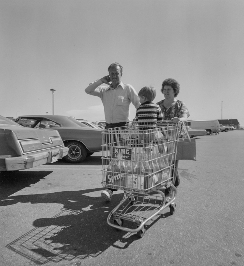 Black-and-white photograph of a man and woman pushing a shopping cart containing a child and cases of Sprite soda through a parking lot filled with cars.