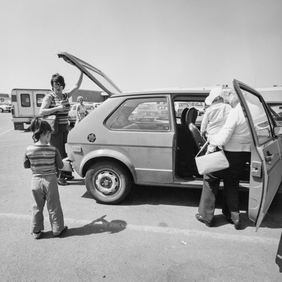 Black-and-white photograph of two women and two children loading items into a Volkswagen hatchback car, a woman wearing sunglasses watches from the background