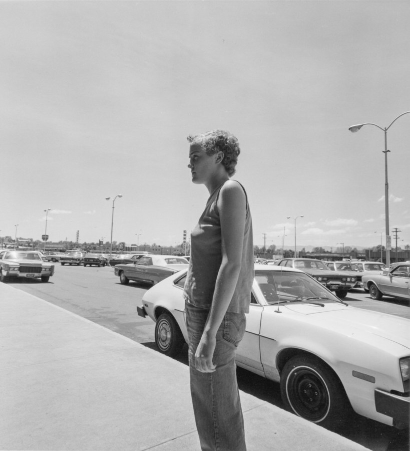 Black-and-white photograph of a person looking to the left in front of a parking lot filled with cars
