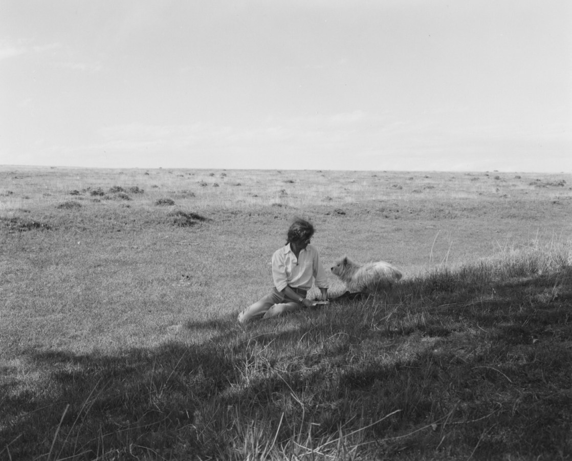A black and white photograph of a woman and a small white dog sitting in a field.