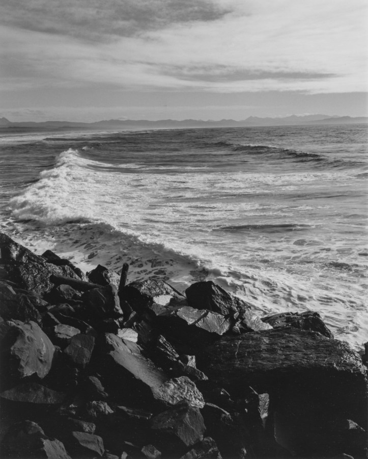 Black-and-white photograph of a wave breaking against a rocky jetty