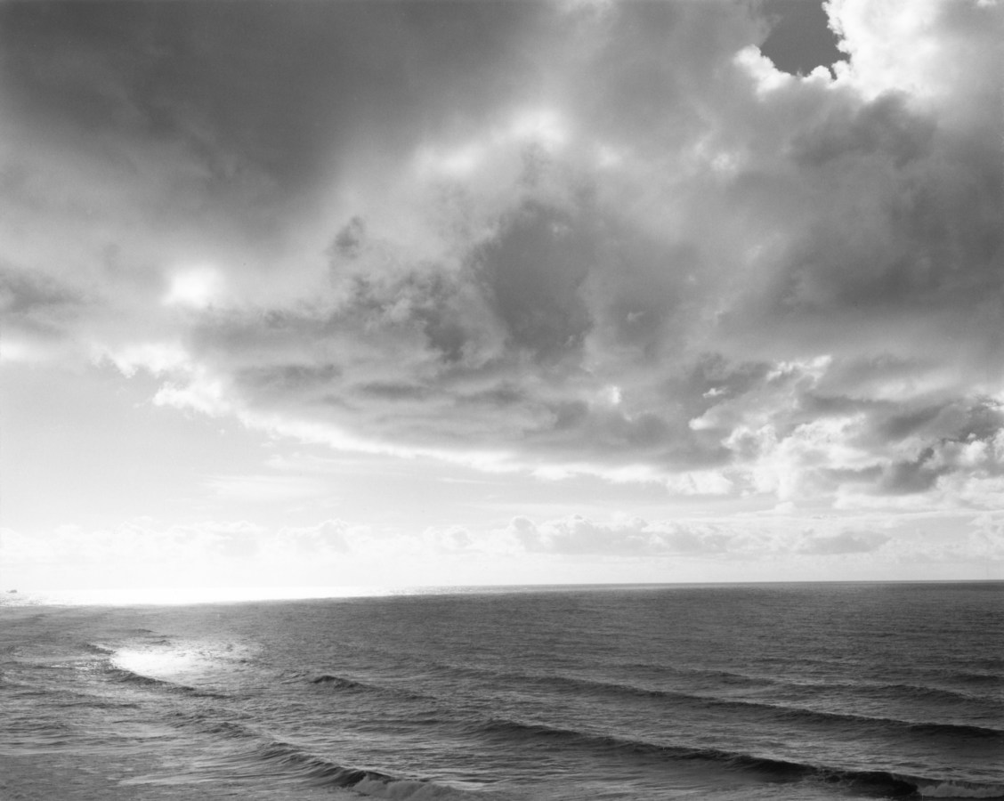 Black-and-white photograph of the ocean with small waves and a cloudy sky with sunlight shining through the clouds
